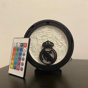 Puede incluir: Un emblema deportivo iluminado en blanco y negro. El emblema es circular con una corona y un logotipo del equipo. Un mando a distancia con m&uacute;ltiples opciones de color se encuentra a la izquierda del emblema.