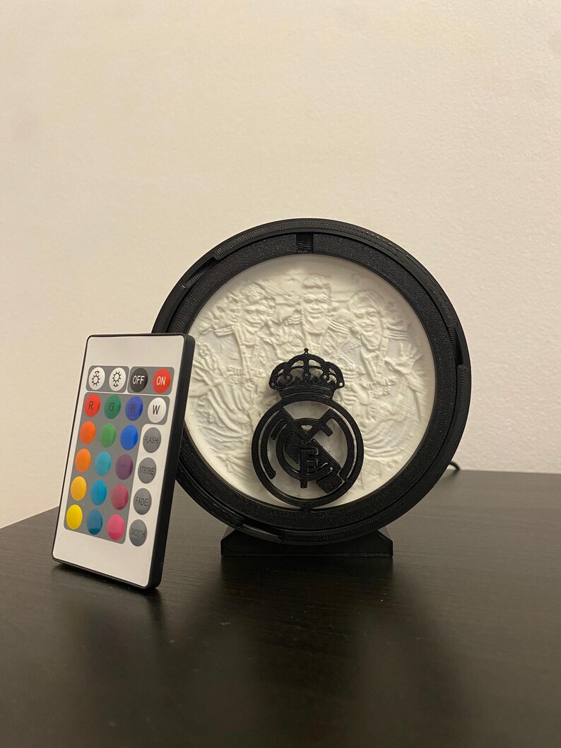 Puede incluir: Un emblema deportivo iluminado en blanco y negro. El emblema es circular con una corona y un logotipo del equipo. Un mando a distancia con m&uacute;ltiples opciones de color se encuentra a la izquierda del emblema.