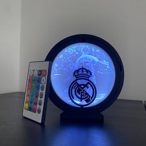 Puede incluir: Una luz LED negra y circular con el logotipo de un equipo deportivo y una imagen de un jugador. Se incluye un mando a distancia con opciones de color. La imagen en el interior es de un futbolista con el nombre "ARDA GULER" y el n&uacute;mero "15".
