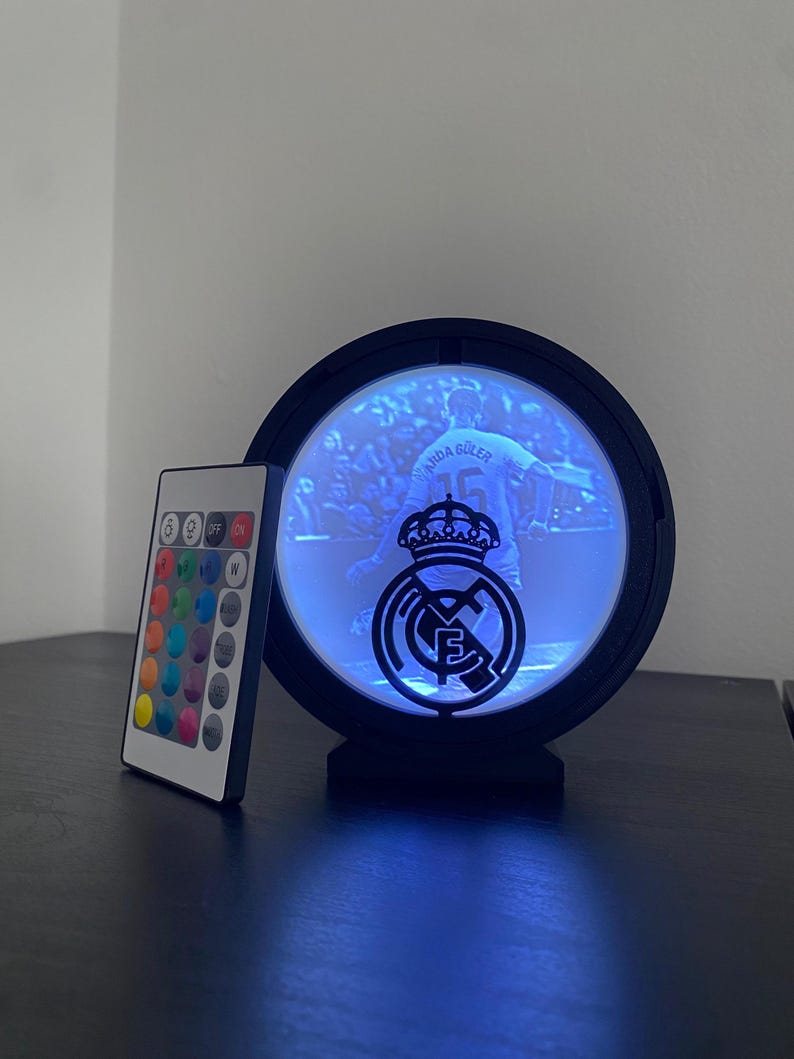 Puede incluir: Una luz LED negra y circular con el logotipo de un equipo deportivo y una imagen de un jugador. Se incluye un mando a distancia con opciones de color. La imagen en el interior es de un futbolista con el nombre "ARDA GULER" y el n&uacute;mero "15".