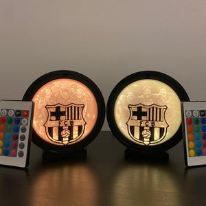 Puede incluir: Dos l&aacute;mparas circulares iluminadas con el logo del FC Barcelona y una foto del equipo. Cada l&aacute;mpara tiene un marco negro y un mando a distancia. Las l&aacute;mparas emiten un brillo c&aacute;lido, mostrando el emblema y los jugadores del equipo.