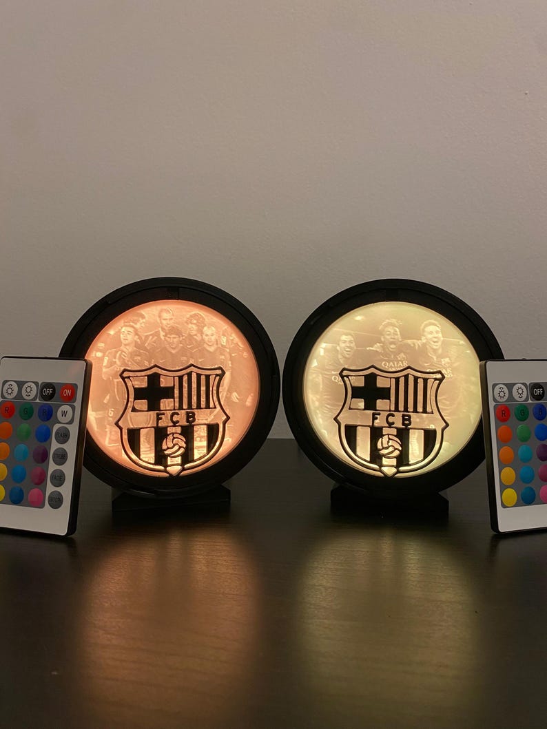 Puede incluir: Dos l&aacute;mparas circulares iluminadas con el logo del FC Barcelona y una foto del equipo. Cada l&aacute;mpara tiene un marco negro y un mando a distancia. Las l&aacute;mparas emiten un brillo c&aacute;lido, mostrando el emblema y los jugadores del equipo.