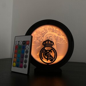 Puede incluir: Una l&aacute;mpara LED negra y circular con una imagen de jugador de f&uacute;tbol y el logotipo del equipo. La l&aacute;mpara tiene un mando a distancia con opciones de color. La l&aacute;mpara est&aacute; sobre una superficie oscura.