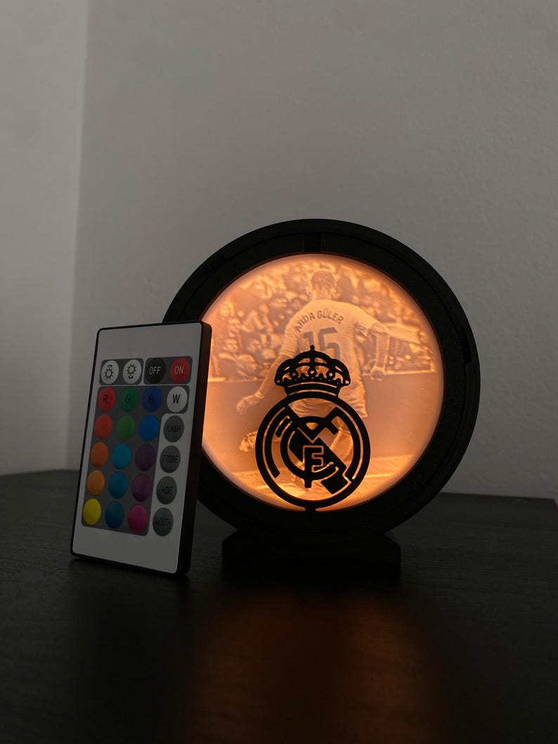 Puede incluir: Una l&aacute;mpara LED negra y circular con una imagen de jugador de f&uacute;tbol y el logotipo del equipo. La l&aacute;mpara tiene un mando a distancia con opciones de color. La l&aacute;mpara est&aacute; sobre una superficie oscura.