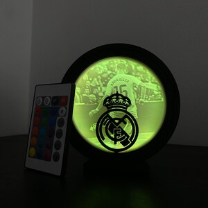 Puede incluir: Una imagen redonda iluminada con marco negro que muestra a un futbolista y el logo del Real Madrid. La imagen brilla en verde, y a la izquierda hay un mando a distancia con opciones de color. El nombre y el n&uacute;mero del jugador son visibles.