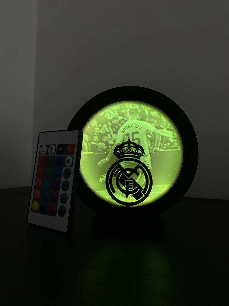 Puede incluir: Una imagen redonda iluminada con marco negro que muestra a un futbolista y el logo del Real Madrid. La imagen brilla en verde, y a la izquierda hay un mando a distancia con opciones de color. El nombre y el n&uacute;mero del jugador son visibles.