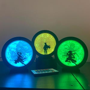 Op de afbeelding: Drie ronde LED-lampen met zwarte frames, elk met een ander silhouet van een anime-personage. De linkerlamp geeft blauw licht, de middelste geel en de rechterlamp groen. Een kleine rechthoekige afstandsbediening ligt aan de voorzijde.