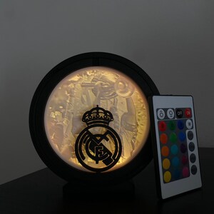 Puede incluir: L&aacute;mpara LED circular negra con el logotipo del Real Madrid. La l&aacute;mpara tiene una imagen detallada en el interior y viene con un mando a distancia para ajustar el color. La l&aacute;mpara est&aacute; dise&ntilde;ada para colocarse sobre una superficie plana.