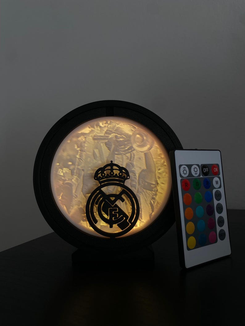Puede incluir: L&aacute;mpara LED circular negra con el logotipo del Real Madrid. La l&aacute;mpara tiene una imagen detallada en el interior y viene con un mando a distancia para ajustar el color. La l&aacute;mpara est&aacute; dise&ntilde;ada para colocarse sobre una superficie plana.