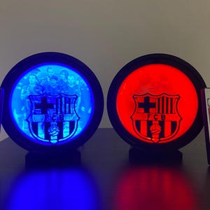 Puede incluir: Dos l&aacute;mparas redondas iluminadas con el logotipo del FC Barcelona. Una l&aacute;mpara brilla en azul, mostrando una foto del equipo, mientras que la otra brilla en rojo. Cada l&aacute;mpara tiene un mando a distancia para ajustar el color. Las l&aacute;mparas est&aacute;n sobre una superficie oscura.
