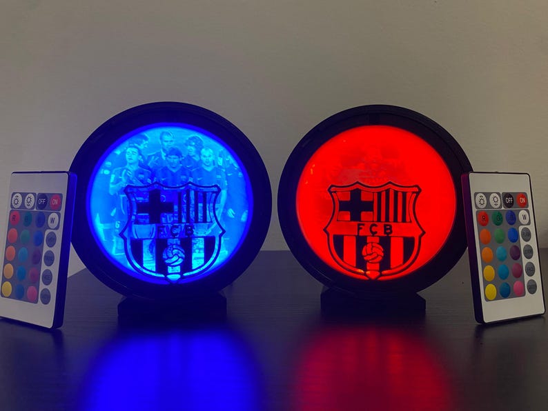 Puede incluir: Dos l&aacute;mparas redondas iluminadas con el logotipo del FC Barcelona. Una l&aacute;mpara brilla en azul, mostrando una foto del equipo, mientras que la otra brilla en rojo. Cada l&aacute;mpara tiene un mando a distancia para ajustar el color. Las l&aacute;mparas est&aacute;n sobre una superficie oscura.