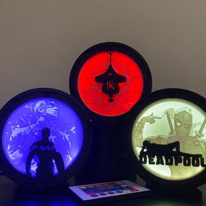 Può includere: Tre lampade LED circolari con cornici nere che presentano sagome di supereroi. La lampada a sinistra brilla di blu con Captain America, quella in alto di rosso con Spider-Man e quella a destra di bianco con Deadpool e il testo DEADPOOL.