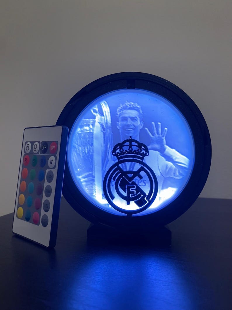 Puede incluir: Una l&aacute;mpara LED redonda con marco negro, que muestra a un jugador de f&uacute;tbol y el logotipo de un equipo. La luz emite un brillo azul, y se ve un mando a distancia con opciones de color. El logotipo del equipo est&aacute; en el centro.