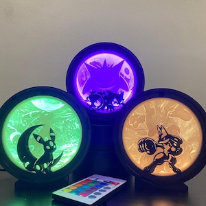 Könnte beinhalten: Drei kreisförmige LED-Lampen mit schwarzem Rahmen, die jeweils die Silhouette einer anderen Figur zeigen. Eine leuchtet grün mit einer Kreatur auf einer Mondsichel, eine violett mit einer stacheligen Kreatur und eine warmgelb mit einer Kämpferfigur.