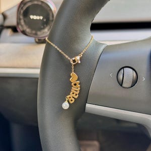 Puede incluir: Un collar de tono dorado con una cadena fina y un colgante que incluye un pequeño corazón, el nombre BELLA en letras mayúsculas y una cuenta de perla blanca que cuelga en la parte inferior. La joya se muestra sobre un volante oscuro.