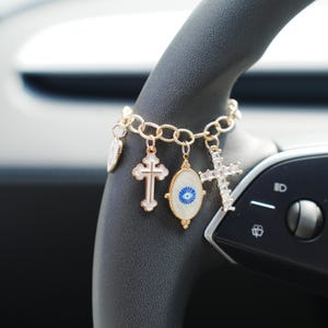Könnte beinhalten: Ein goldfarbenes Bettelarmband mit vier verschiedenen Anhängern: einem kleinen runden Anhänger, einem weißen Emaille-Kreuz mit Goldrand, einem ovalen Schutzaugen-Anhänger in Blau und Weiß sowie einem mit klaren Kristallen besetzten Kreuz.
