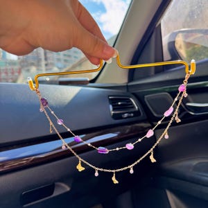 Puede incluir: Un colgante para espejo retrovisor de coche en metal dorado con dos cadenas delicadas adornadas con cuentas de piedras preciosas pulidas en morado y rosa, pequeños cristales transparentes y diminutos dijes de mariposa dorados, sujeto a un gancho.