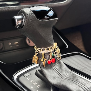 Può includere: Un accessorio a catena in metallo dorato avvolto attorno a una leva del cambio per auto in pelle nera. La catena presenta diversi ciondoli pendenti, tra cui una lettera R dorata, una stella a quattro punte, due ciliegie rosse e un fiocco.