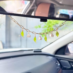 May include: A gold-toned metal chain hanging from a rearview mirror, featuring alternating charms of small green leaves and white bell-shaped flowers. The chain consists of elongated oval links, with each charm attached to a separate link along the strand.