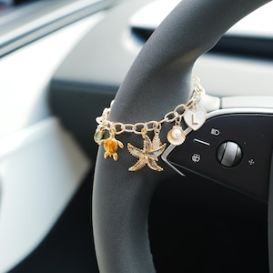 Könnte beinhalten: Ein goldfarbenes Kettenarmband mit mehreren Anhängern im Ozean-Design, darunter eine Meeresschildkröte, ein Seestern, eine Muschel mit einer Perle und ein weißes herzförmiges Element mit dem Buchstaben L, befestigt an ovalen Kettengliedern.