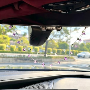 May include: A car rearview mirror hanging ornament featuring two delicate silver-toned chains adorned with small, polished purple amethyst and pale green fluorite gemstone beads, suspended from the mirror mount in a graceful, draped arc.