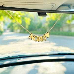 May include: A gold-toned metal chain hanging from a rearview mirror, featuring individual letter beads that spell out the name NORA, followed by a small, solid gold-toned heart charm. The letters and heart are smooth, rounded, and reflective.