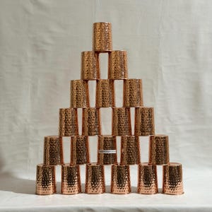 Könnte beinhalten: Eine Pyramide aus kupferfarbenen, gehämmerten Metallbechern. Die Becher sind in einer gestapelten Formation angeordnet, wobei jede Schicht die Anzahl der Becher verringert. Die Becher haben eine strukturierte Oberfläche und eine zylindrische Form.