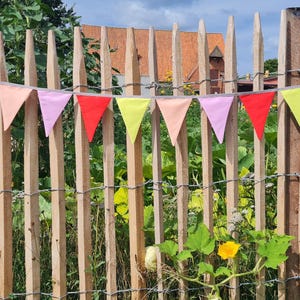 Könnte beinhalten: Eine Wimpelkette mit dreieckigen Flaggen in den abwechselnden Farben Hellrosa, leuchtendes Rot und Limettengelb. Die Wimpel sind an einer dünnen Schnur befestigt und horizontal vor einem vertikalen Holzzaun im Außenbereich angebracht.