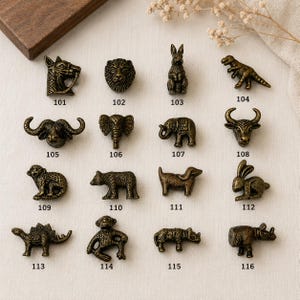 Op de afbeelding: Een verzameling van zestien antieke messingkleurige metalen kastknoppen in diervorm, elk gelabeld met een driecijferig nummer van 101 tot 116. De figuren omvatten een paard, leeuw, konijn, dinosaurus, buffel, olifant, stier, aap, beer, hond en neushoorn.