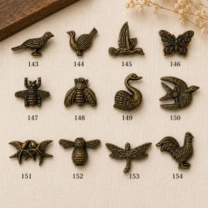 May include: A collection of twelve antique bronze-toned metal charms shaped like animals and insects, labeled with numbers 143 through 154. The set includes figures of birds, a butterfly, a bee, a swan, a dragonfly, and a rooster, each with detailed textures.