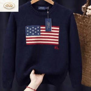 Peut inclure: Pull-over bleu marine avec un grand drapeau américain sur le devant. Le drapeau présente des étoiles blanches sur un champ bleu et des rayures rouges et blanches. Les lettres "RL" sont brodées en rouge. Le pull a un col rond et des manches longues.