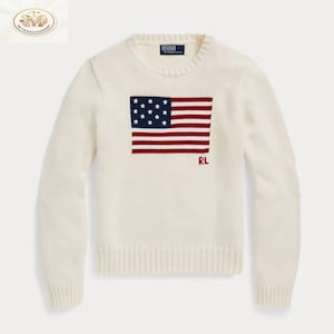 May include: Cream-colored sweater featuring a red, white, and blue American flag design on the front. The sweater has long sleeves, a crew neck, and ribbed cuffs and hem. The letters "RL" are embroidered below the flag.