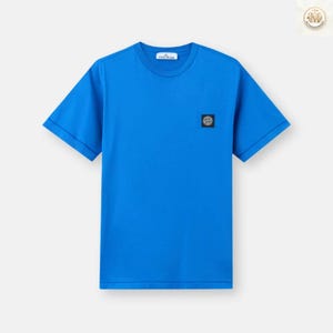 Peut inclure: Un t-shirt bleu vif à manches courtes. Un petit écusson carré avec un emblème circulaire est apposé sur la poitrine gauche. Le t-shirt a un col rond et un design classique, adapté aux tenues décontractées.