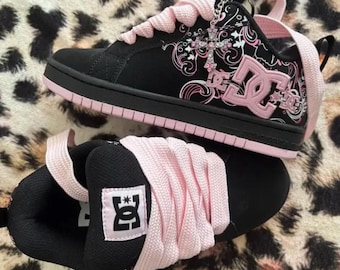 May include: Black and light pink DC skate sneakers featuring thick pink laces, a prominent DC logo on the side, and a silver cross charm attached to the eyelets. The shoes have a black sole and a black upper with white and pink graphic patterns.
