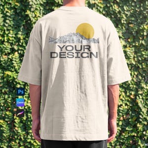 May include: A beige oversized t-shirt featuring a graphic on the back. The graphic displays a black and white mountain range illustration topped with a solid yellow circle representing the sun. Below the mountains, the text reads YOUR DESIGN.