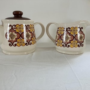 May include: A vintage cream-colored teapot with a matching creamer. Both pieces feature a repeating floral pattern in shades of brown, yellow, and cream. The teapot has a brown lid and a curved spout. The creamer has a handle and a pouring spout.