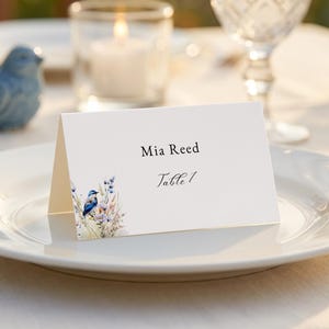 May include: A white place card with the name "Mia Reed" and "Table 1" printed in elegant script. The card features a watercolor illustration of a blue bird and floral accents. The card is placed on a white plate.