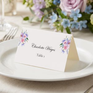 May include: A folded place card with floral watercolor accents in pink, purple, and blue. The card reads "Charlotte Hayes" and "Table 1" in elegant script. The card is set on a white plate, with flowers in the background.