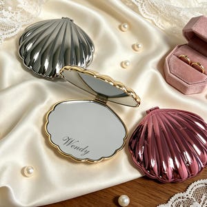 Op de afbeelding: Drie schelpvormige compacte spiegels in zilver, goud en metallic roze. Eén gouden spiegel staat open en toont de naam Wendy gegraveerd in het glas. Een klein roze fluwelen ringdoosje met twee gouden ringen staat ernaast, omringd door parels.