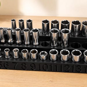 May include: A black socket organiser with two tiers, holding a variety of shiny, silver sockets. The sizes are marked on the front from 4 to 15. The sockets are arranged neatly, suggesting a tool set for automotive or mechanical work.