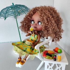 May include: A doll with voluminous curly brown hair, wearing a green dress with a yellow pineapple print, platform shoes, and a small bag. The doll holds a tiny glass of yellow liquid while sitting on a white chair under a teal wire umbrella.