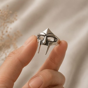 Puede incluir: Un anillo de metal plateado con un diseño de máscara estilizada que presenta una frente puntiaguda, aberturas horizontales para los ojos y dos barras verticales que se extienden hacia abajo desde la nariz, con acabado pulido y detalles oscuros.