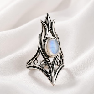Op de afbeelding: Een zilveren ring met een ovale maansteen cabochon, gezet in een puntig, architecturaal metalen frame. De ringband vertoont gedetailleerde uitsparingen en krulwerk, met een gepolijste afwerking die de blauwe iriserende glans van de steen benadrukt.