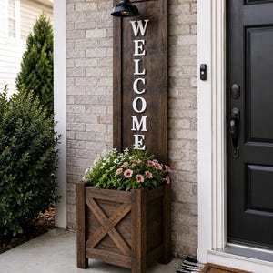May include: A wooden welcome sign with a built-in planter box. The sign displays the word "WELCOME" in white letters and is topped with a black light fixture. The planter is filled with pink and white flowers.