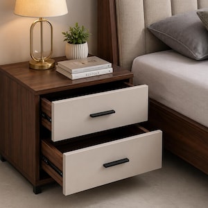 May include: A two-drawer bedside table with a dark brown wooden frame and light beige drawer fronts. The drawers have black handles. A gold lamp, a small potted plant, and books are on top. The table is next to a bed.