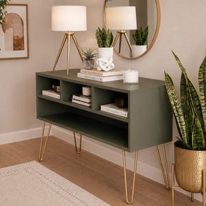 May include: A sage green console table with gold hairpin legs, holding books, candles, and decorative items. Two white lamps with gold tripods sit on top. A large potted plant and a gold-toned mirror are also visible.