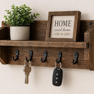 May include: A rustic wooden key holder with a shelf, featuring a small potted plant and a framed sign that reads "HOME sweet home." Below the shelf are five black hooks, with keys and a car remote hanging from some.