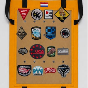 Op de afbeelding: Een felgeel rechthoekig displaybord met zwarte riemen en een kleine Nederlandse vlag aan de bovenkant. Het bord heeft een beige middenpaneel met zestien diverse geborduurde patches, waaronder ontwerpen met tekst zoals WHY ME, I BB, UCA en FRAGILE.