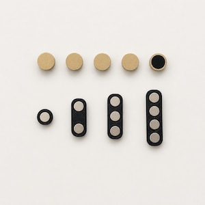 May include: A collection of magnetic fasteners including four circular tan discs, one circular tan disc with a black centre, and four black rectangular magnetic strips featuring one, two, three, or four silver circular magnets embedded in each piece.