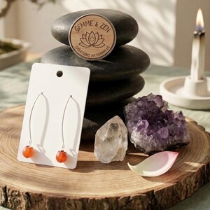 Peut inclure: Une paire de boucles d'oreilles pendantes argentées avec des perles orange rondes sur un support blanc. Le support porte le texte Gemme & Zen Creations Artisanales avec un motif de lotus. Les boucles sont près d'un cristal et d'une améthyste.